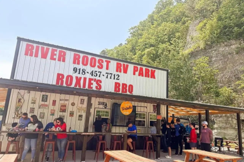 A busy outdoor BBQ spot at River Roost RV Park, with people gathered under a shaded area and a scenic backdrop.