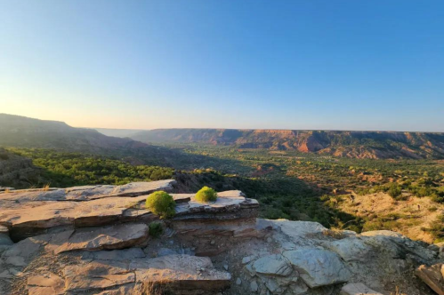 A scenic view of a canyon landscape with rocky cliffs and lush greenery under a clear blue sky.