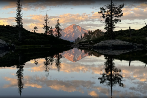 A serene landscape featuring a mountain reflected in a calm lake, surrounded by trees and a colorful sky at sunset.
