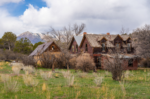 Abandoned houses surrounded by overgrown grass and trees, with snow-capped mountains in the background under a cloudy sky.