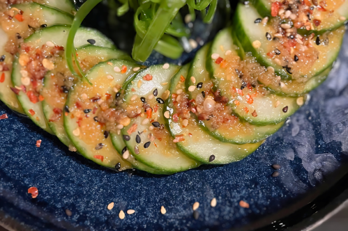 Sliced cucumbers arranged in a circular pattern, topped with sauce, sesame seeds, and spices on a blue plate.