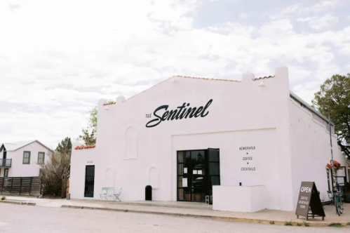 A white building with "The Sentinel" sign, featuring large windows and an open sign outside, set against a cloudy sky.