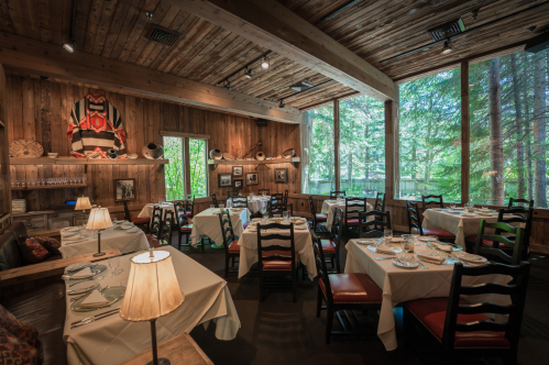 Cozy restaurant interior with wooden decor, tables set for dining, and large windows overlooking a forested area.