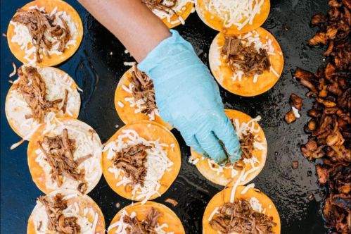A hand in a blue glove adds shredded meat to cheese-topped tortillas on a griddle.