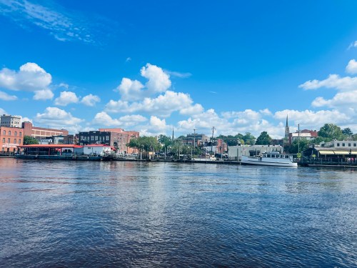 A scenic view of a river with buildings and boats along the shore under a blue sky with fluffy clouds.