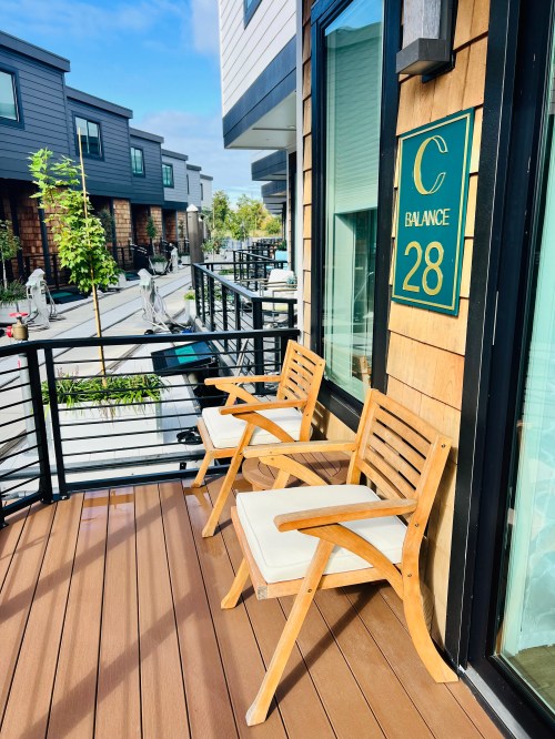 Two wooden chairs with cushions on a balcony, next to a sign that reads "C Balance 28," overlooking a modern neighborhood.
