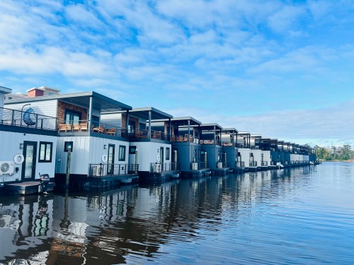 A row of modern houseboats lined up along a calm waterway under a blue sky with scattered clouds.
