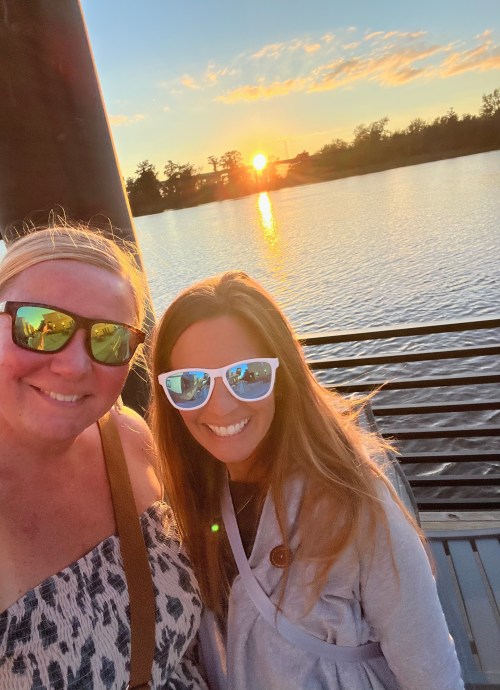 Two women wearing sunglasses smile for a selfie by a lake at sunset, with trees and a colorful sky in the background.