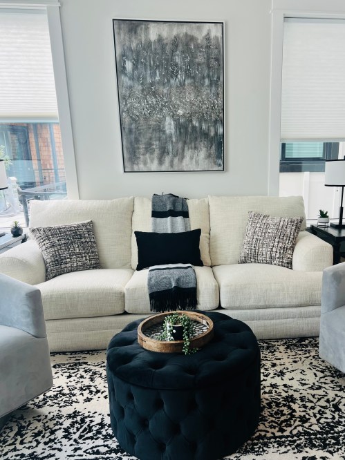A cozy living room with a light sofa, patterned pillows, a round black ottoman, and a large abstract wall art piece.