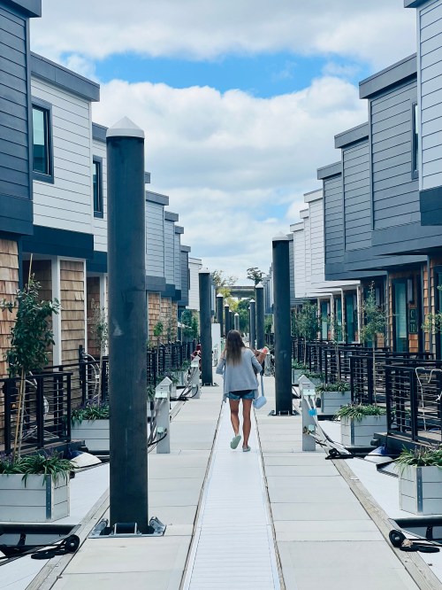 A person walks down a modern pathway lined with buildings and greenery under a partly cloudy sky.
