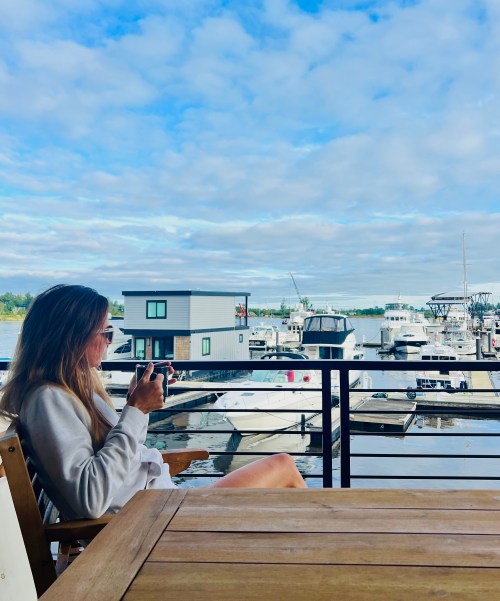 A woman sits on a deck, sipping a drink, with boats and a cloudy sky in the background.