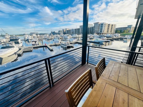 A marina view with boats docked, reflecting clouds and buildings, seen from a wooden deck with chairs.