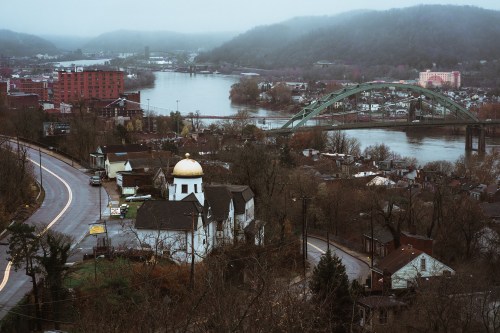 A misty view of a river town with a bridge, buildings, and a church with a golden dome in the foreground.