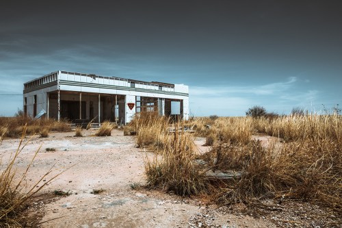 Abandoned building surrounded by tall grass and a clear sky, with a weathered facade and empty surroundings.