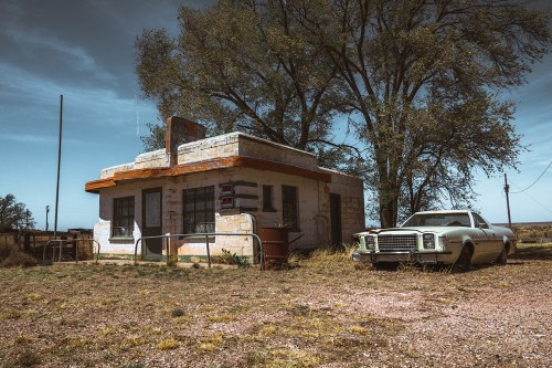 Abandoned building with a vintage car parked outside, surrounded by overgrown grass and trees under a clear blue sky.