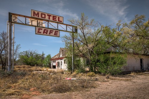 Abandoned motel and café with a weathered sign, surrounded by overgrown vegetation and a clear blue sky.