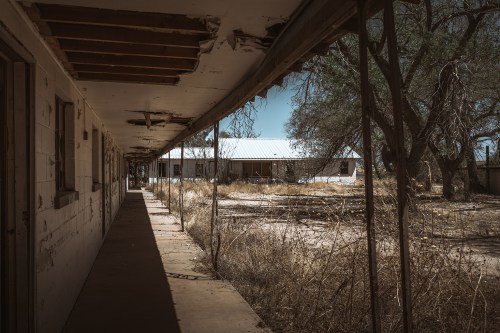 Abandoned building corridor with peeling paint, overgrown grass, and a distant structure under a clear blue sky.