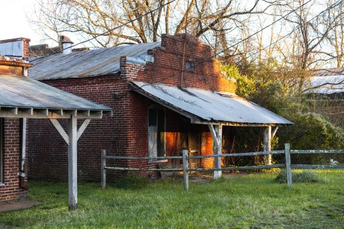 A weathered brick building with a metal roof, surrounded by grass and a wooden fence, illuminated by soft morning light.