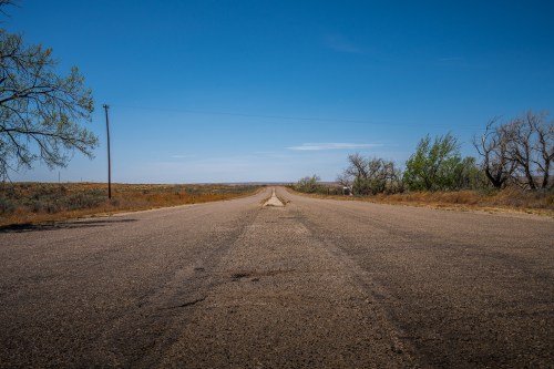 A long, empty road stretches into the distance under a clear blue sky, flanked by sparse vegetation and trees.