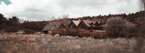 An abandoned farmhouse with a vintage car, surrounded by dry grass and mountains under a cloudy sky.