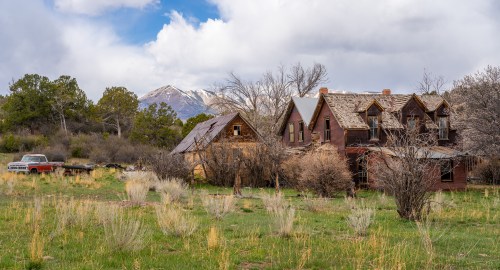 Abandoned houses with overgrown vegetation, set against a backdrop of mountains and cloudy skies.