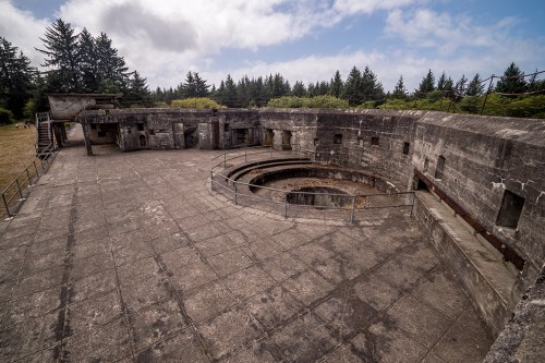 An abandoned concrete military structure with a circular opening, surrounded by trees and a cloudy sky.
