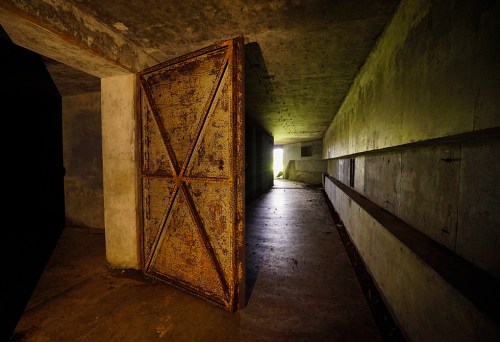 An open rusty door leads into a dimly lit, abandoned concrete corridor.