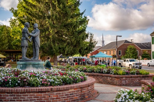 A statue of two figures shaking hands in a park, surrounded by flowers and a market in the background.