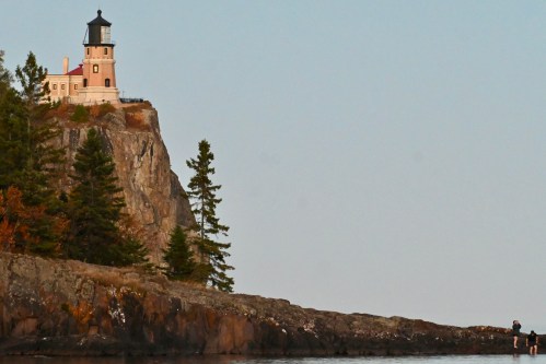 A lighthouse atop a rocky cliff, surrounded by trees, with a person fishing on the shore nearby.