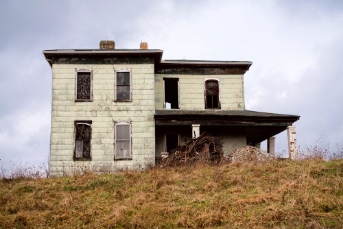 Abandoned green house on a hill, with broken windows and overgrown grass under a cloudy sky.