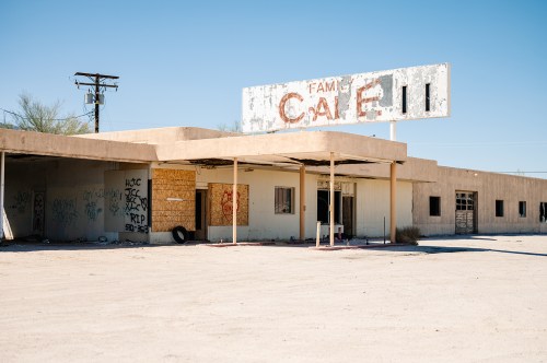 Abandoned café building with a weathered sign, surrounded by graffiti and a barren landscape under a clear blue sky.