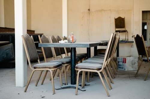 A vacant dining area with a large table surrounded by empty chairs and a single glass jar on the table.