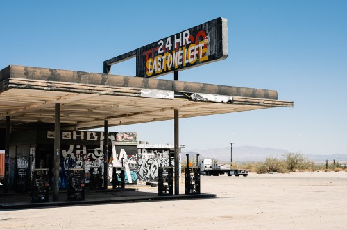 Abandoned gas station with graffiti, featuring a "24 HR" sign against a clear blue sky and desert landscape.