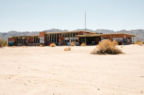 Abandoned building in a desert landscape, covered in graffiti, with mountains in the background and sparse vegetation.