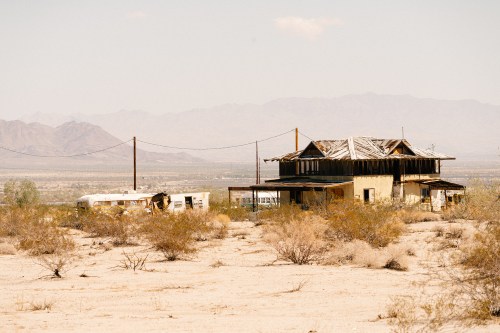 Abandoned house and trailer in a desert landscape, with mountains in the background and sparse vegetation.