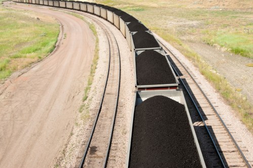 A long train of coal cars curves along a dirt path, with green grass and hills in the background.