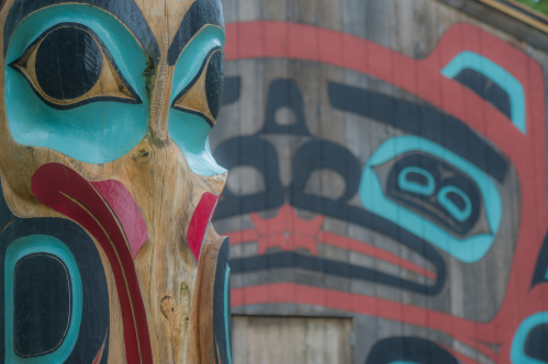 A close-up of a colorful totem pole with intricate designs, set against a wooden background featuring additional artwork.