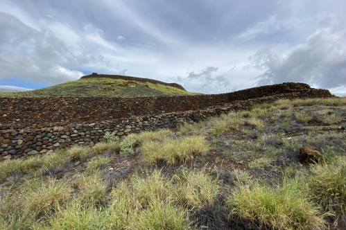 Stone walls curve through grassy terrain, leading to a hill under a cloudy sky.