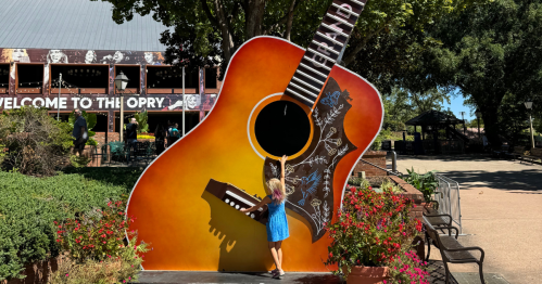 A child in a blue dress reaches for a giant guitar sculpture surrounded by flowers at a venue entrance.