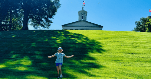 A child in a cap plays on a grassy hill with a historic building and flags in the background under a clear blue sky.