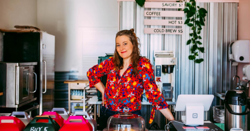 A smiling barista in a colorful shirt stands behind a coffee shop counter with drinks and pastries on display.