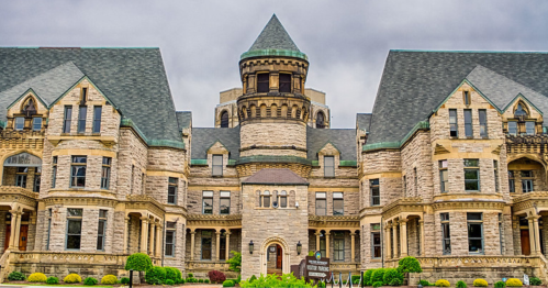 Historic stone building with a turret, intricate architecture, and a green roof, set against a cloudy sky.