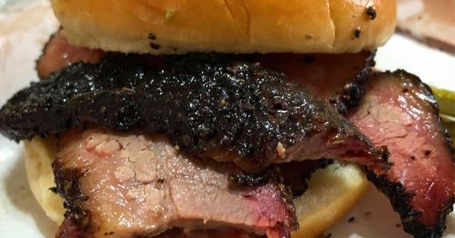 Close-up of a sandwich with layers of smoked brisket on a soft bun, showcasing a dark, crispy crust.