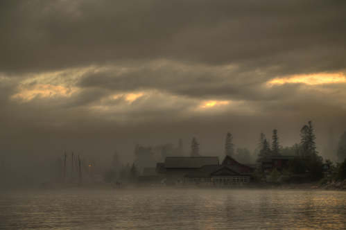Misty lake scene at dawn with silhouettes of trees, buildings, and boats under a cloudy sky.