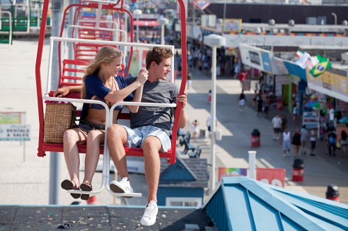 A couple enjoys a scenic ride on a chairlift above a bustling boardwalk filled with shops and people.