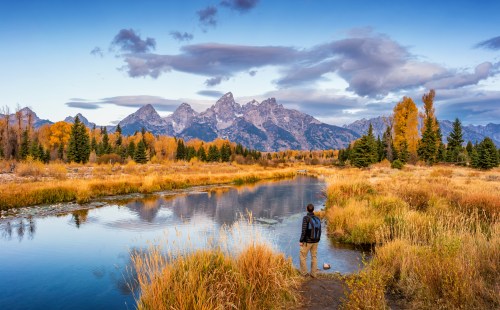 A person stands by a river, surrounded by autumn foliage and mountains under a blue sky with clouds.