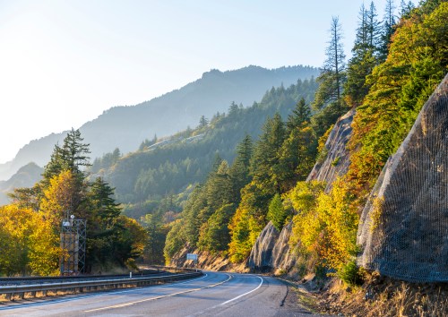 A winding road through a mountainous landscape with trees and autumn foliage under a soft morning light.