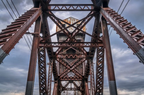 View from below a large, rusted metal bridge structure against a cloudy sky, showcasing intricate beams and supports.