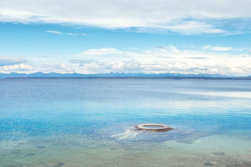 A serene lake with clear blue water, a rocky formation in the foreground, and distant mountains under a cloudy sky.