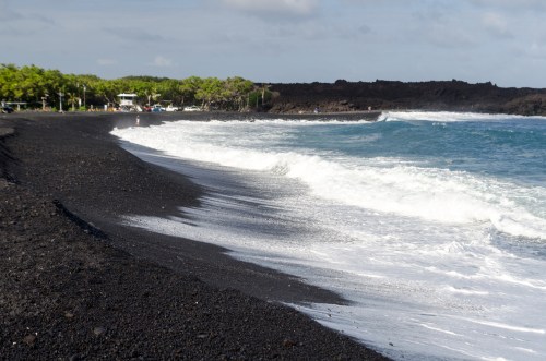A scenic beach with black sand, gentle waves, and lush greenery in the background under a partly cloudy sky.